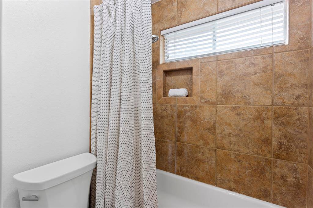 The separate shower space features a tiled shower with a curtain, a built-in shelf, and a window with blinds for natural light.