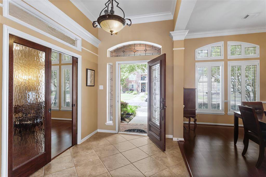 This inviting entryway features a stylish chandelier, elegant wooden doors with decorative glass, and abundant natural light from large windows with plantation shutters. The tiled floor and neutral walls create a warm, welcoming atmosphere, with views of a lush front yard.