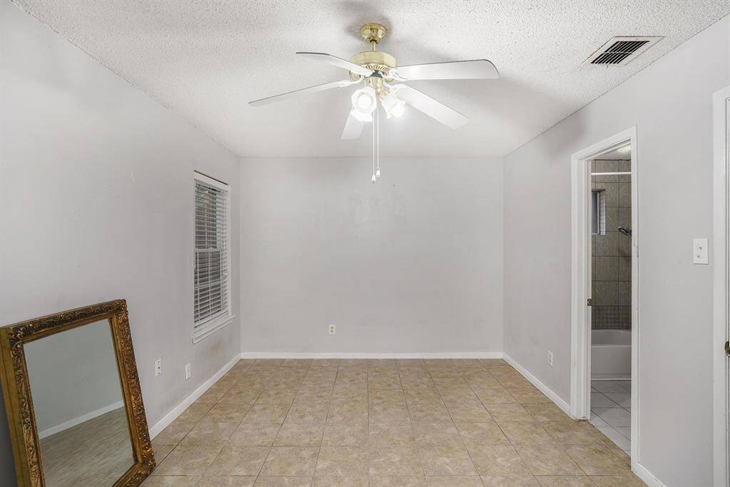 This room features neutral-colored walls and tile flooring, complemented by a ceiling fan for comfort. The space includes a window for natural light and an attached bathroom, making it functional and versatile.