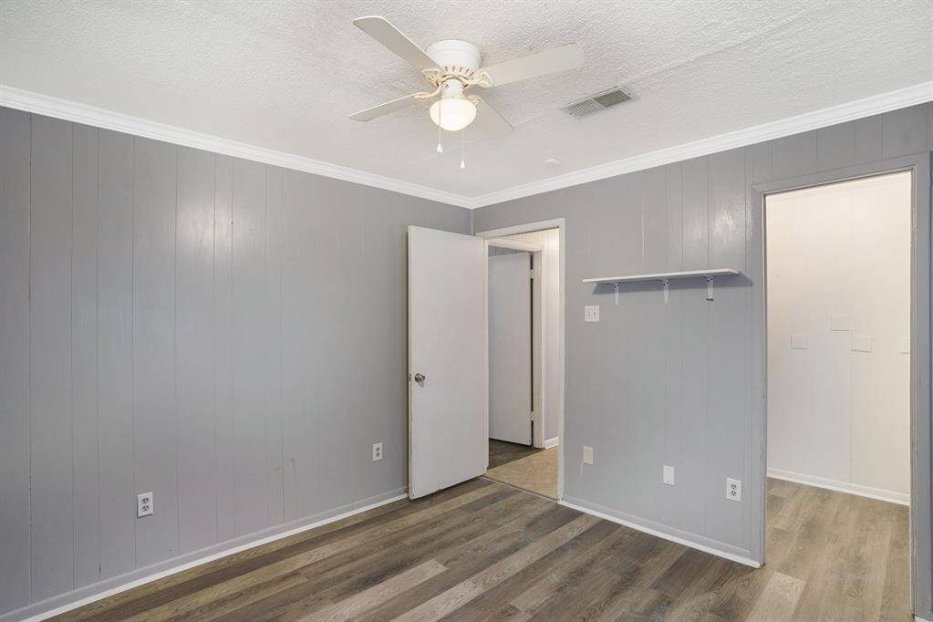 This room features gray paneled walls, a ceiling fan, and laminate wood flooring. It includes a small wall shelf and is well-lit with natural light through an open doorway. Suitable for a bedroom or office.
