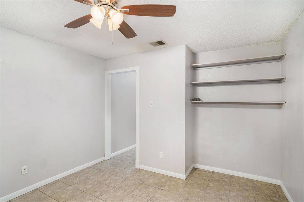 This room features neutral-colored walls and tile flooring, with a ceiling fan for comfort. It includes built-in shelves for added storage and a doorway leading to an adjoining space.