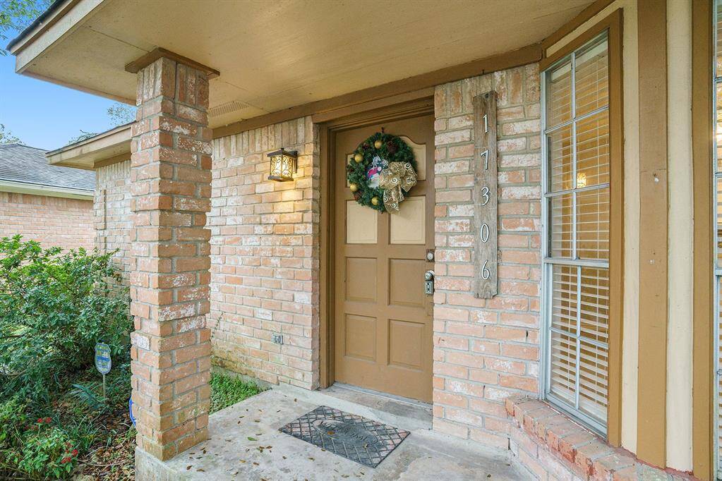 This welcoming home entrance features a classic brick exterior, a wooden front door adorned with a festive wreath, and a cozy covered porch with a charming light fixture. A large window with shutters adds natural light to the inviting space.