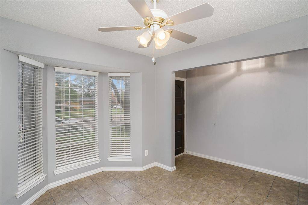 Bright dining area with large bay windows, neutral gray walls, and tiled flooring. Features a ceiling fan and opens into an adjacent space, creating an inviting atmosphere.