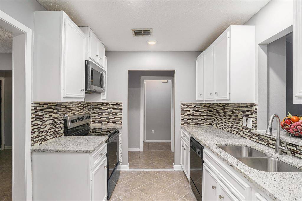 This modern kitchen features white cabinetry, granite countertops, and a stylish mosaic tile backsplash. It includes stainless steel appliances, a double sink, and a cozy, well-lit atmosphere with a neutral color palette.