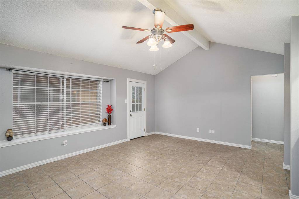 This room features a neutral color palette with a vaulted ceiling and a ceiling fan. It has tiled flooring, a large window with blinds, and a door leading outside. The space is bright and airy, ideal for a cozy living area.