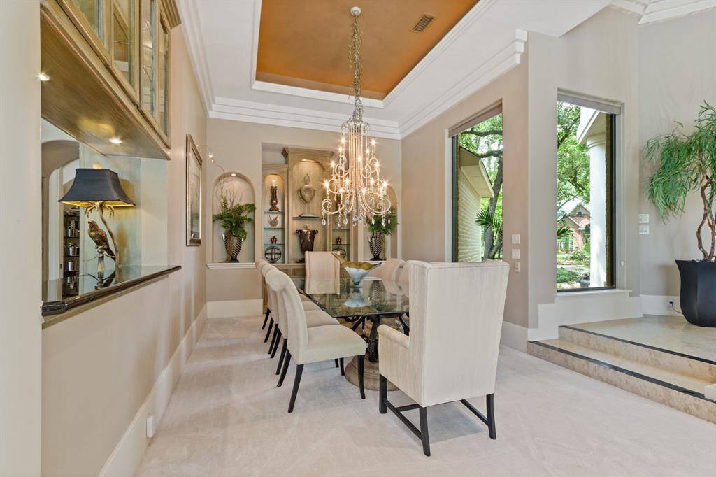 The photo showcases an elegant spacious dining room highlighted by a large ornate chandelier, crown molding, and high decorative ceiling. Built-in lighted display shelves for your art pieces, and large windows offering ample natural light. The neutral color palette create a luxurious atmosphere.