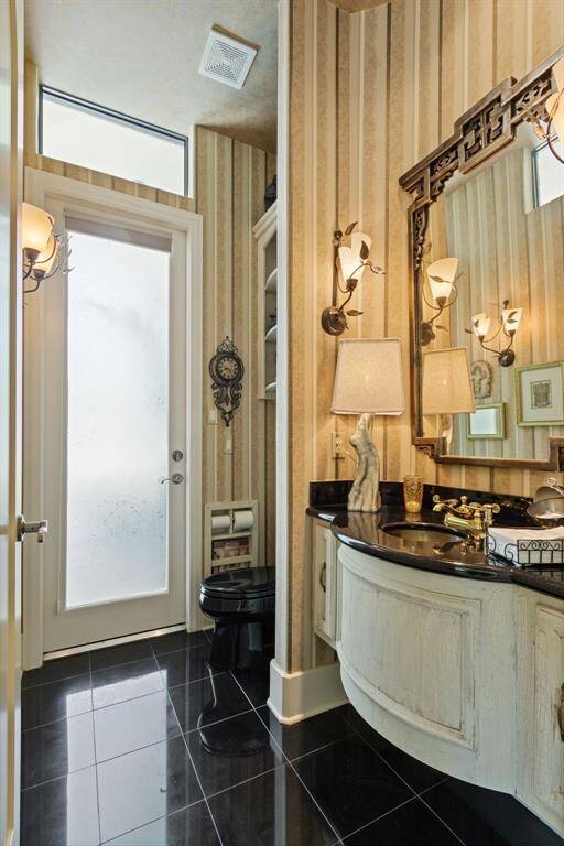 Chic pool bathroom with elegant striped wallpaper and a black countertop. Features a stylish black toilet, ornate mirror, and ample lighting. Frosted glass door provides natural light and privacy. Perfect blend of modern and classic design.