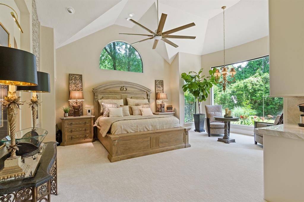 This spacious bedroom features high ceilings, a large arched window, and modern ceiling fan. It has a cozy, elegant design and a seating area by the expansive window overlooking lush greenery, providing plenty of natural light.