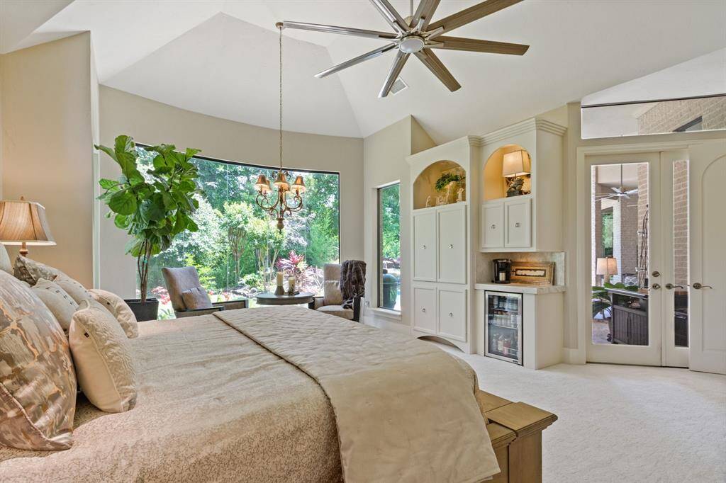 This spacious bedroom features large windows with lush garden views, a vaulted ceiling and elegant lighting. It includes built-in cabinetry, a place for a coffee bar, juice/wine fridge, and French doors leading to an outdoor space, offering a perfect blend of comfort and style.