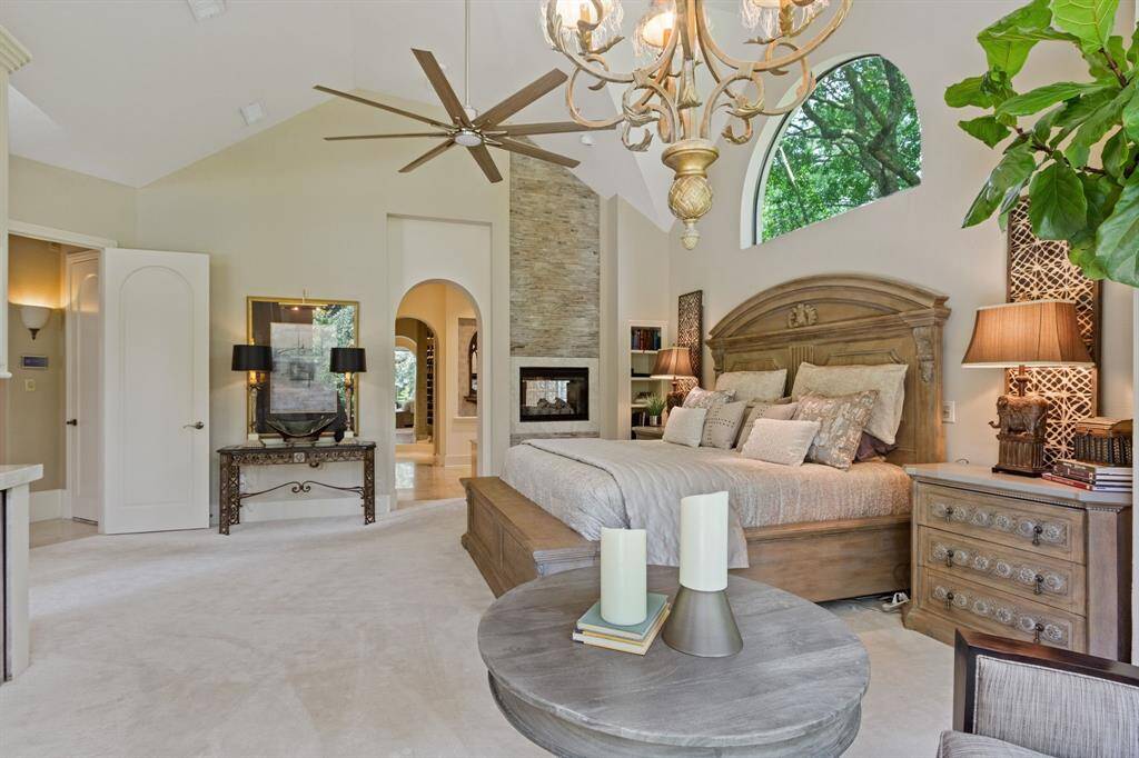 This spacious bedroom features a high ceiling with a modern fan and elegant chandelier. It has a cozy see-through fireplace, with a carved wooded accent wall, large arched window offering natural light. The room provides a luxurious and serene atmosphere.