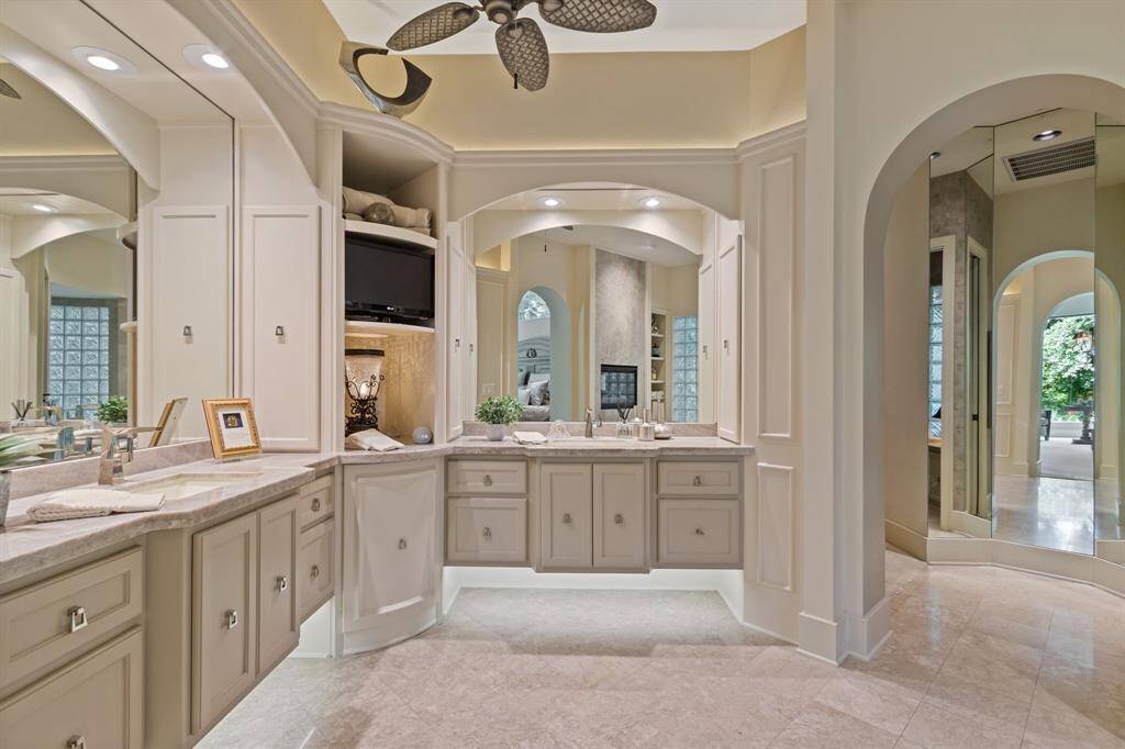 This elegant expansive bathroom features dual vanities with ample storage, gorgeous Taj Mahal Quartzite counters and a ceiling fan. The neutral color palette and arched doorways complement the spacious and well-lit design, creating a luxurious atmosphere.