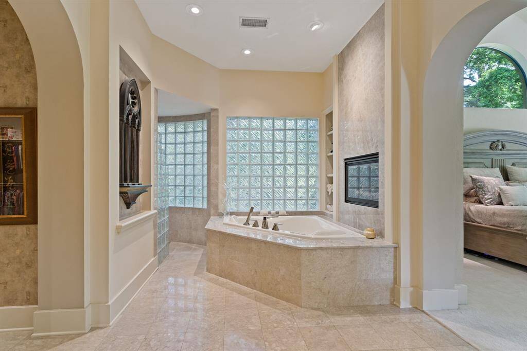 This photo showcases a luxurious bathroom with a custom hydrotherapy tub with champagne bubble jets. Elegant tile work and glass block windows, that provides natural light. The space includes a fireplace for added comfort, and an open archway leads to a cozy bedroom, creating a seamless flow between the rooms.