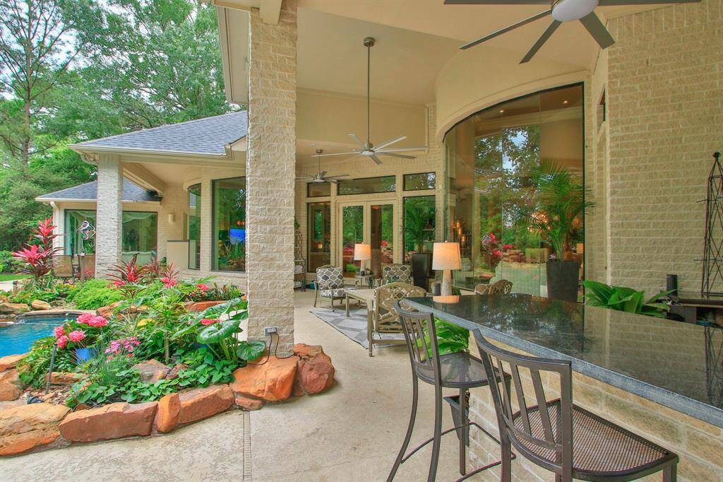 This photo showcases a beautiful outdoor living space with a covered patio featuring ceiling fans and ample space for your outdoor furniture. There's a stone bar for entertaining, and large windows provide a view into the lush garden and pool area, surrounded by vibrant landscaping. Ideal for relaxation and gatherings.