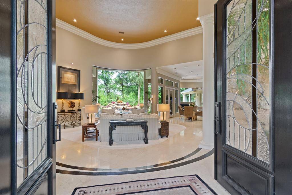 This elegant entryway showcases double doors leading into a spacious, open-plan living area with large windows offering lush garden views. The interior features high ceilings, polished floors,  creating a welcoming and luxurious atmosphere.