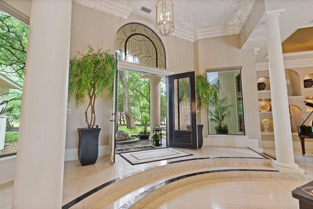 This elegant foyer features high ceilings, large windows, and a grand entrance with double doors. The space is adorned with decorative columns, a stylish chandelier, and built-in shelves showcasing art pieces. Lush greenery outside enhances the inviting atmosphere.