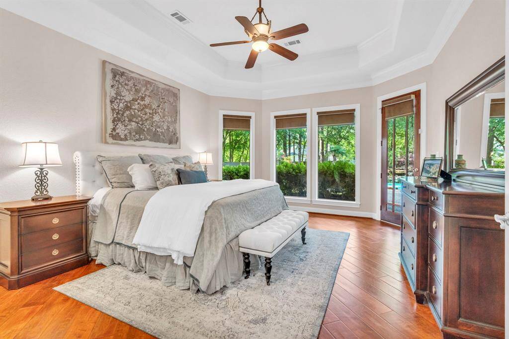 Serene primary suite featuring a tray ceiling with crown molding, rich hardwood floors, and a wall of windows and door with custom coverings overlooking the manicured backyard and pool