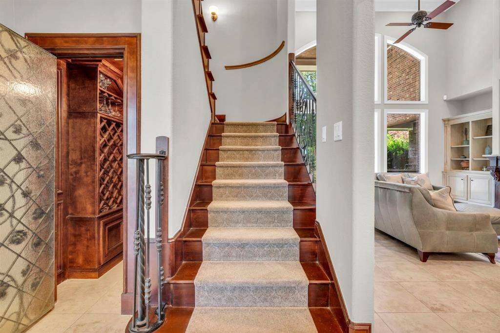 Elegant staircase crowned by wrought-iron balusters and hardwood treads; tucked just off the landing is another view of the custom wine grotto, while soaring windows reveal the living room’s double-height grandeur beyond.