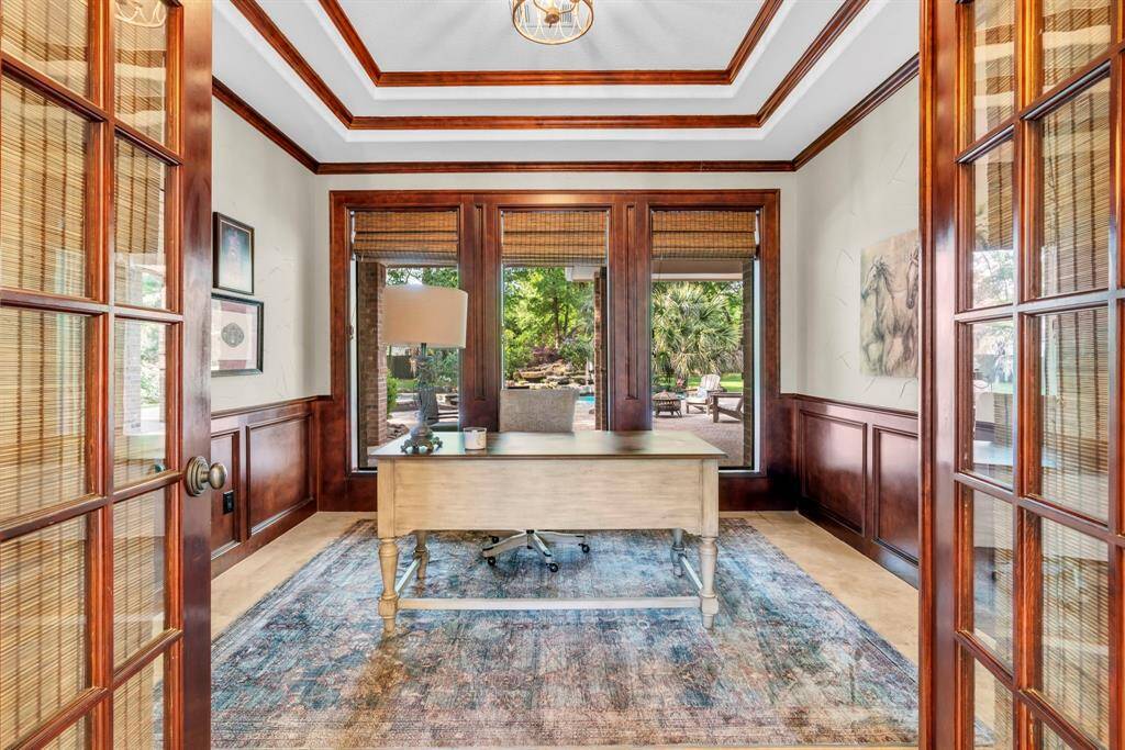 Richly appointed study behind double French doors boasts layered tray ceiling with wood trim, wainscoting, and three picture windows with custom window coverings overlooking the pool—an inspiring space for work or reading.