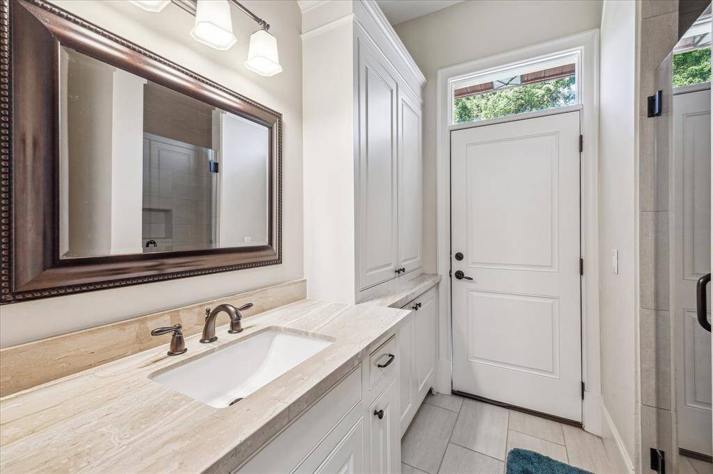 Conveniently located on the first floor, this full bathroom features a walk-in shower, elegant tile flooring, and a spacious vanity with ample storage and stone countertops. A large framed mirror and stylish lighting add warmth and character, while a private exterior door enhances accessibility—perfect for guests or seamless use from the outdoor living area.