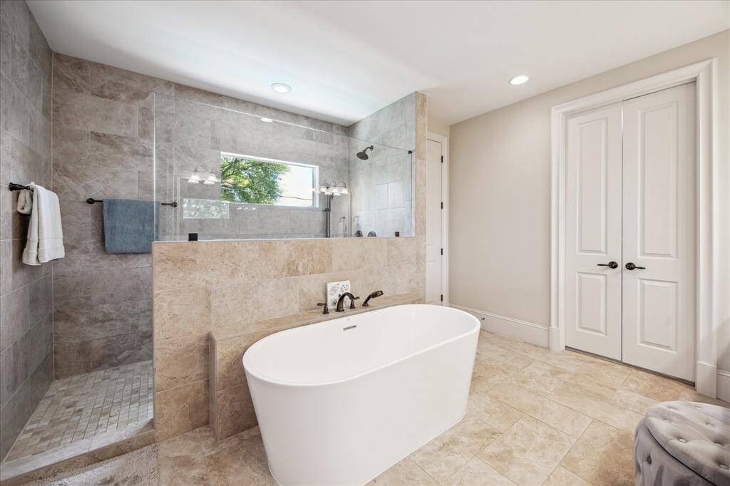Sophisticated and spa-like, the primary bathroom features a sleek freestanding soaking tub set against a backdrop of neutral stone tile. A frameless glass shower with built-in bench and dual shower heads adds both luxury and convenience. Soft natural light filters through the privacy window, while thoughtful finishes and generous space create a serene retreat for daily routines or evening relaxation.