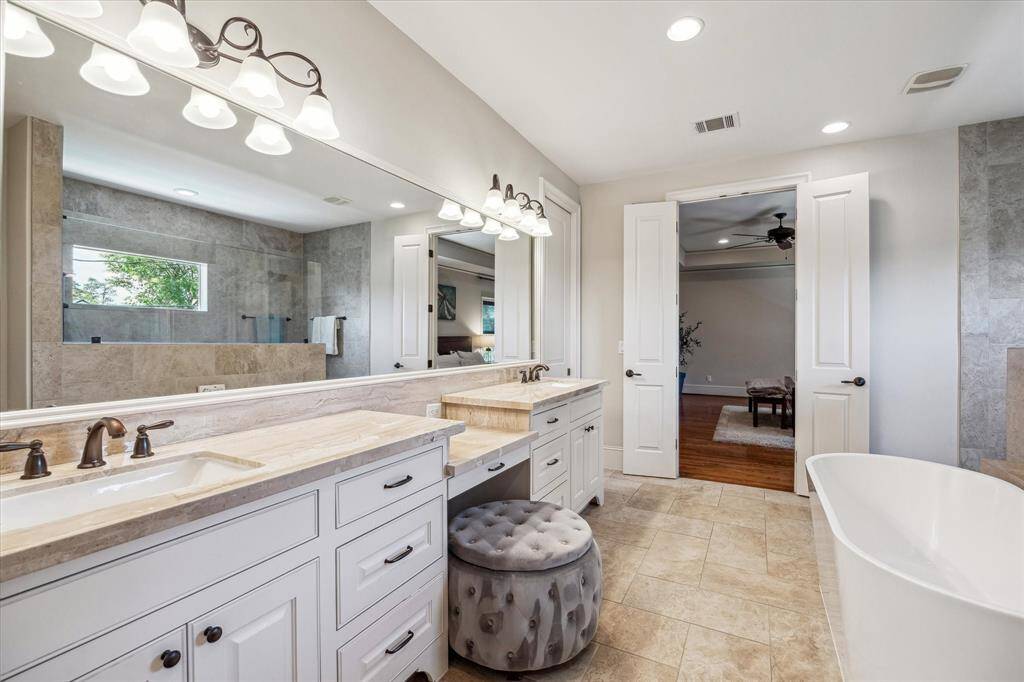 This alternate view showcases the dual vanities with abundant drawer storage and a spacious countertop layout, including a dedicated seated vanity area. A large framed mirror runs the length of the wall, enhancing the room's sense of space and light. Soft neutral tile underfoot continues the spa-like feel, while the open layout offers direct access to the serene primary suite beyond.