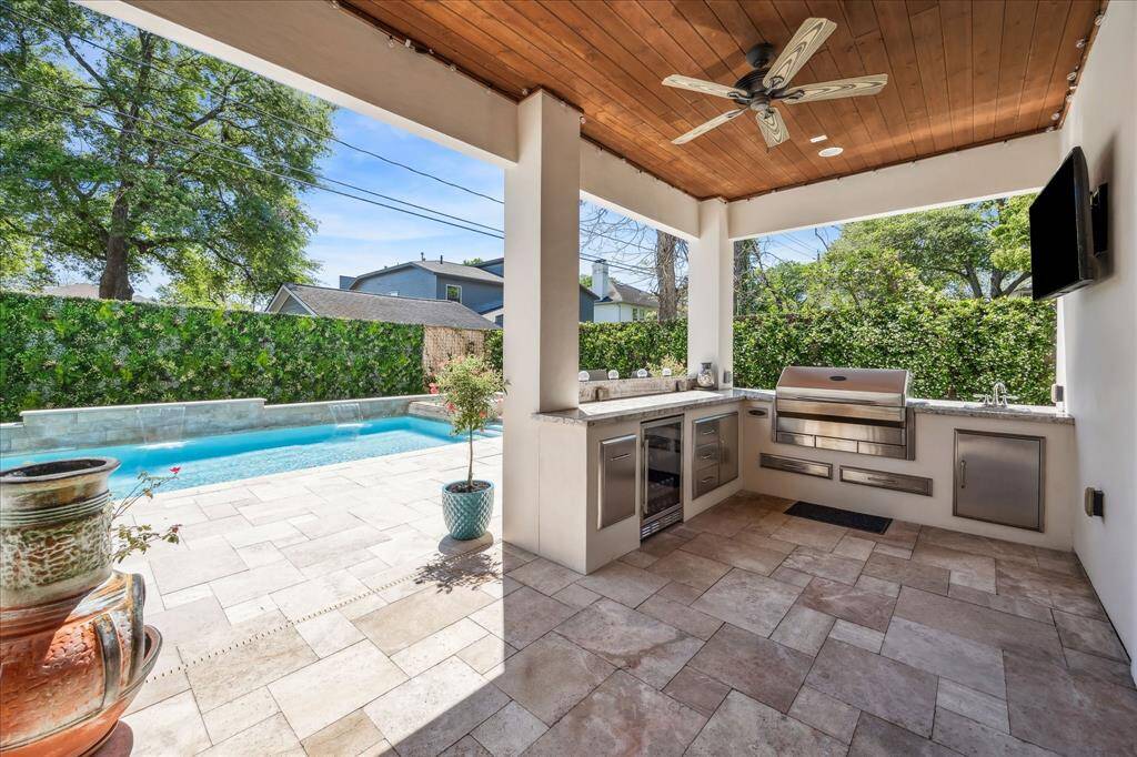 Covered summer kitchen is equipped with a built-in grill, beverage cooler, sink, and prep space—all beneath a rich wood-paneled ceiling with a ceiling fan. Perfectly positioned to overlook the pool, this setup keeps you connected to the fun while prepping a meal or mixing a drink.