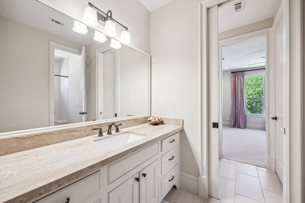 Crisp and well-appointed Jack-and-Jill bathroom featuring a spacious stone countertop, under-mount sink, and bronze fixtures. A large mirror and elegant lighting provide a bright, polished look, while separate access points from each bedroom ensure privacy and ease of use. The adjoining shower area showcases sleek tilework, and ample cabinetry offers practical storage for everyday essentials.