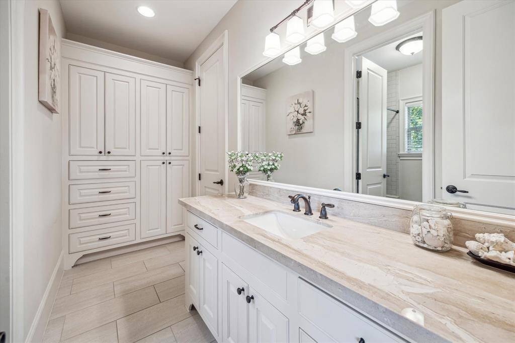 Stylish shared bathroom featuring a long stone-topped vanity with ample cabinet storage and sleek bronze fixtures. Light neutral tile flooring and soft wall tones create a clean, calming aesthetic, while a full wall of built-in cabinetry offers excellent organizational space. Conveniently situated between a bedroom and the game room, this shared bath is as functional as it is refined.