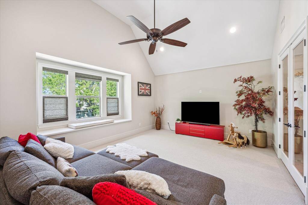 Spacious upstairs game room with vaulted ceilings, plush carpet, and abundant natural light pouring through a charming triple window with a built-in bench seat. A ceiling fan and recessed lighting enhance the comfort and functionality of the space, while double glass doors provide a stylish transition to adjacent rooms. Ideal for movie nights, playtime, or casual lounging.