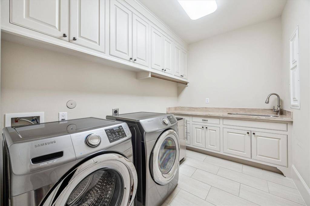 Well-equipped laundry room designed with both style and functionality in mind. Features include upper and lower cabinetry for abundant storage, a stone countertop with built-in sink, and a dedicated hanging rod for convenience. Neutral tile flooring and soft wall tones keep the space bright and clean, making everyday tasks more efficient and organized.