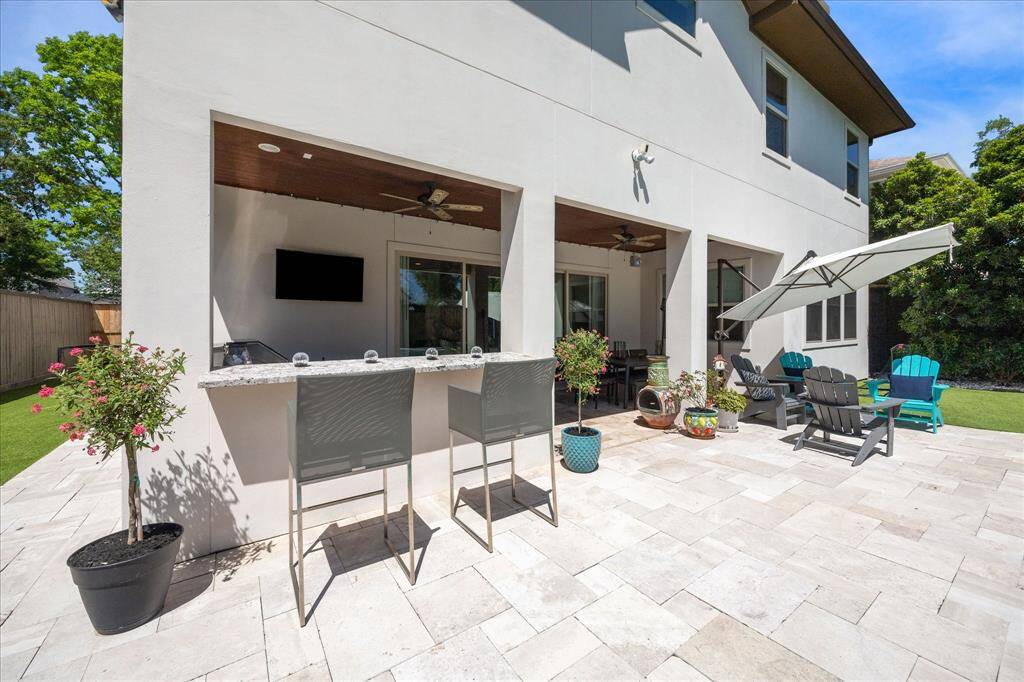 Expansive patio with travertine tile extends from the covered outdoor living area, complete with ceiling fans. A sleek bar with granite countertop offers a seamless setup for serving drinks and casual dining, while the surrounding greenery and open-air layout create a relaxing, resort-style vibe.