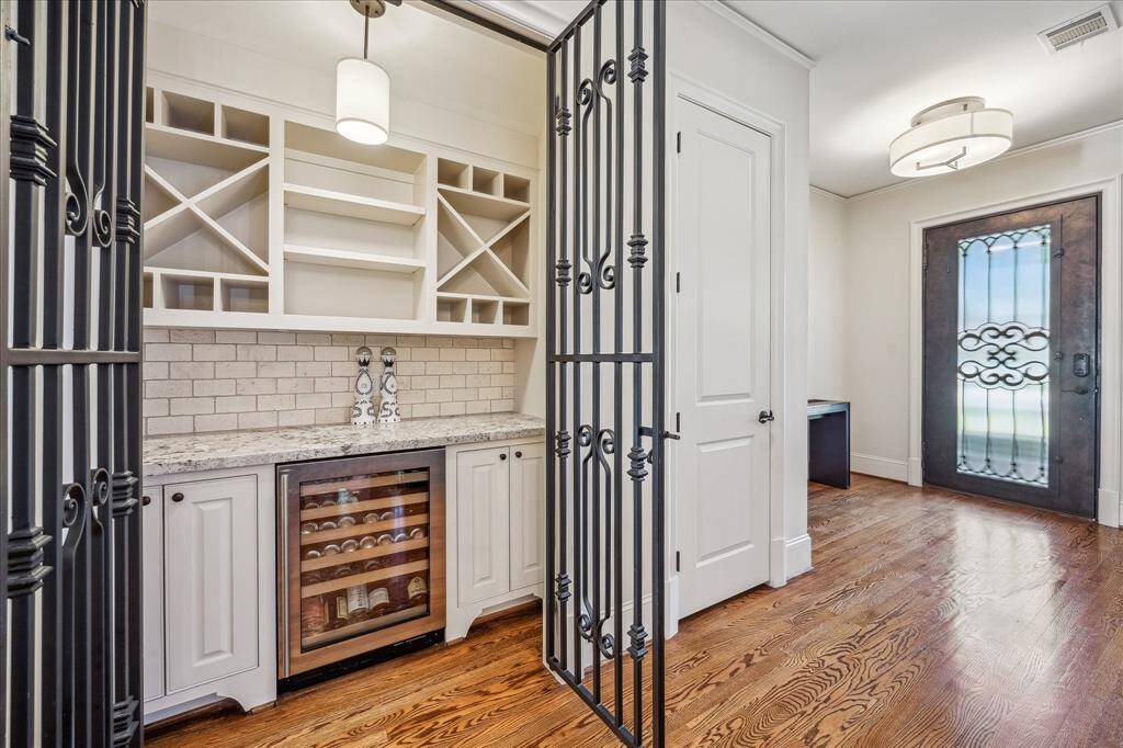 Just off the entry, a beautifully designed wine bar is tucked behind elegant wrought iron gates, offering both style and function. The custom cabinetry includes open shelving for glassware and bottle display, along with a wine fridge and granite countertops. A soft pendant light and classic subway tile backsplash add warmth and character, while the adjacent front door—featuring ornate iron detailing—sets the tone for the thoughtful craftsmanship found throughout the home.