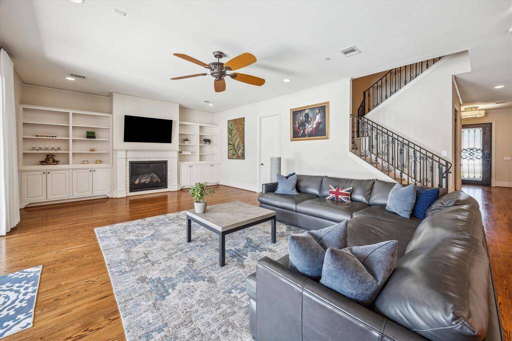 Spacious and light-filled living area anchored by a gas fireplace with a sleek tile surround and flanked by custom built-ins for stylish storage and display. Beautiful hardwood floors and soft neutral walls create a warm, inviting ambiance, while wrought iron stair railings add architectural interest and elegance. Large windows and recessed lighting ensure the space remains bright and airy throughout the day.