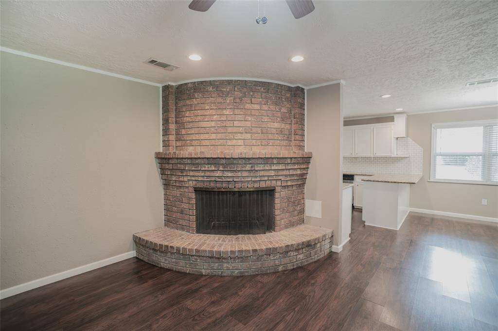 This inviting living space features a unique rounded brick fireplace, adding warmth and character. The open layout connects to a bright kitchen with white cabinetry and a large window, creating a cozy yet spacious atmosphere.