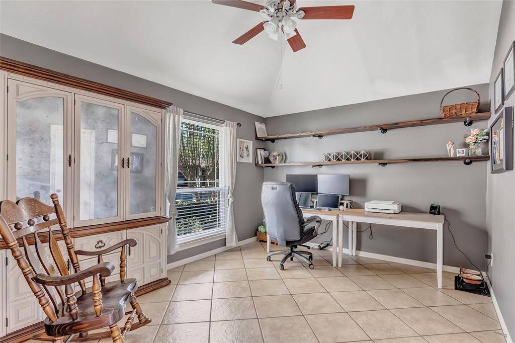 This bright home office features tiled flooring, a ceiling fan, and ample natural light from a large window. It includes a built-in wooden cabinet, floating shelves for storage, and a modern desk setup, creating a functional workspace.