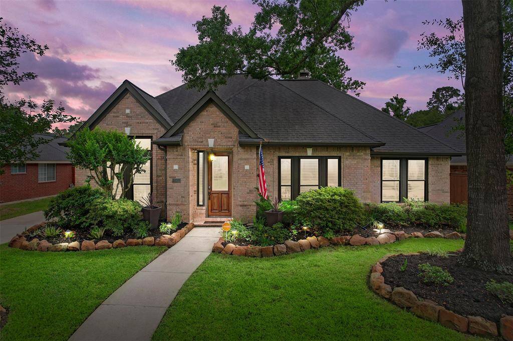 This inviting single-story brick home offers timeless curb appeal with its manicured lawn, mature trees, and soft accent lighting. A welcoming front path leads you through thoughtfully maintained landscaping, all set beneath a sunset sky that sets the tone for peaceful evenings ahead.