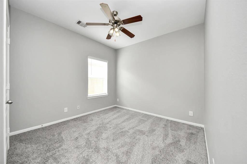 This room features soft gray walls, a plush carpet, and a ceiling fan with lights. A single window allows natural light, creating a bright and cozy space.