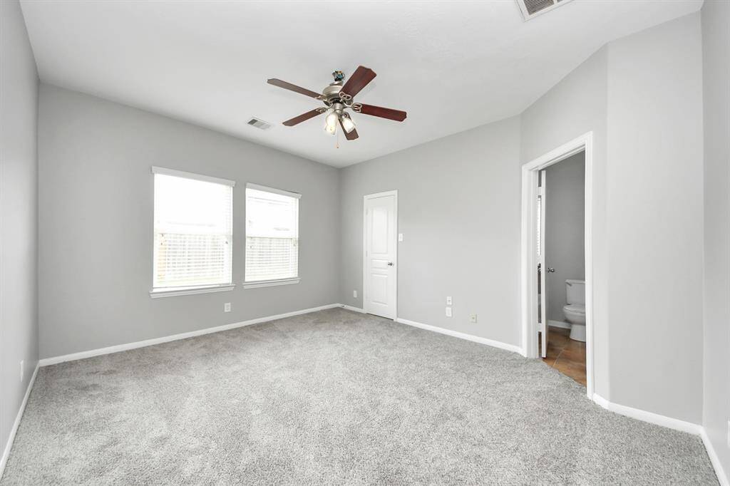This bright primary bedroom features soft gray walls, plush carpeting, a ceiling fan, and two large windows for natural light. There's an attached bathroom, adding convenience and functionality.