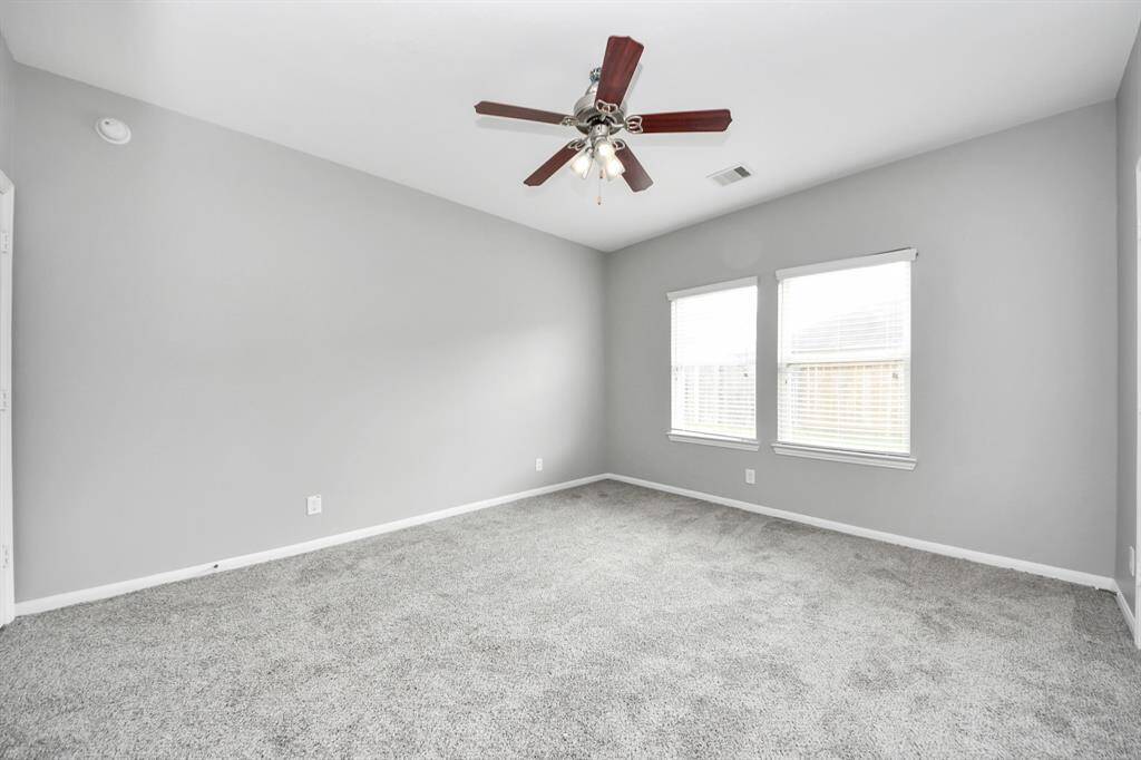 Bright and spacious room with light gray walls, plush carpeting, and a ceiling fan. Two large windows provide ample natural light. Ideal for a bedroom or office space.
