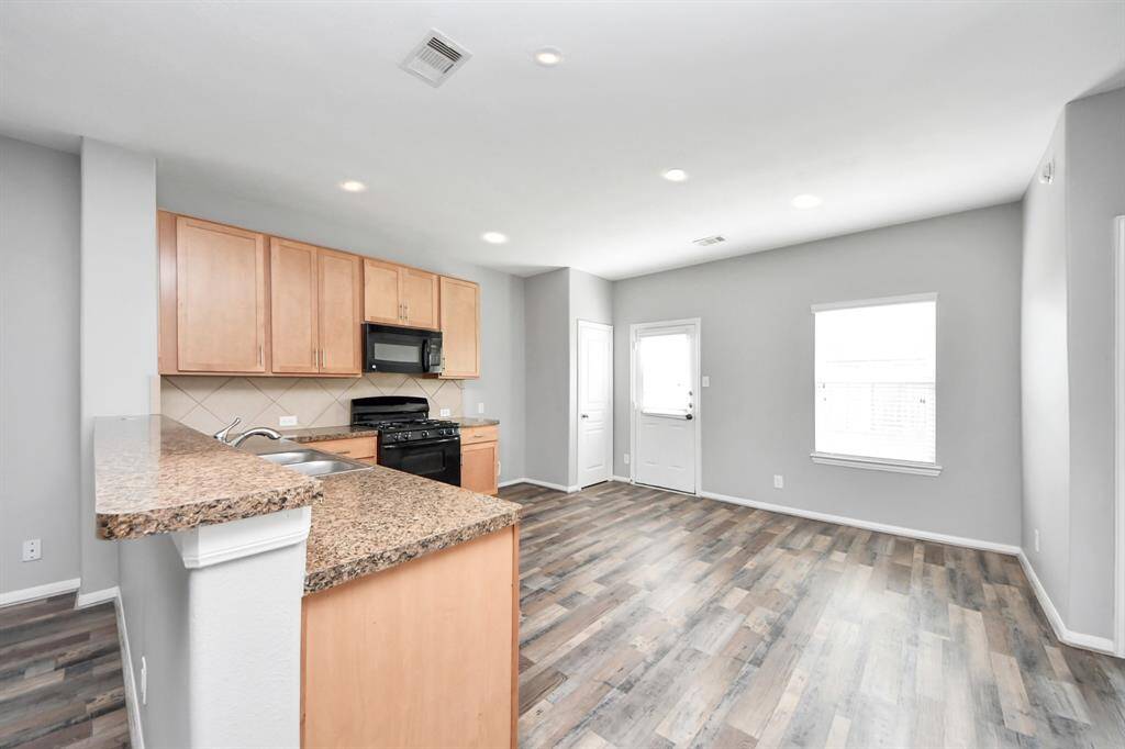 This photo showcases a modern kitchen and dining area with light wood cabinetry, and a tile backsplash. It features a black oven and microwave, hardwood flooring, and ample natural light from a large window and a door leading outside. The space is open and airy, perfect for entertaining.
