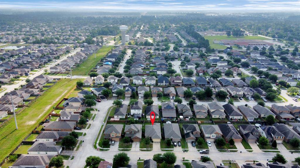This aerial photo showcases a suburban neighborhood with rows of single-family homes. A highlighted house in the center of the image indicates the property for sale. The area is well-developed, with tree-lined streets and a nearby park or recreational field visible in the background.