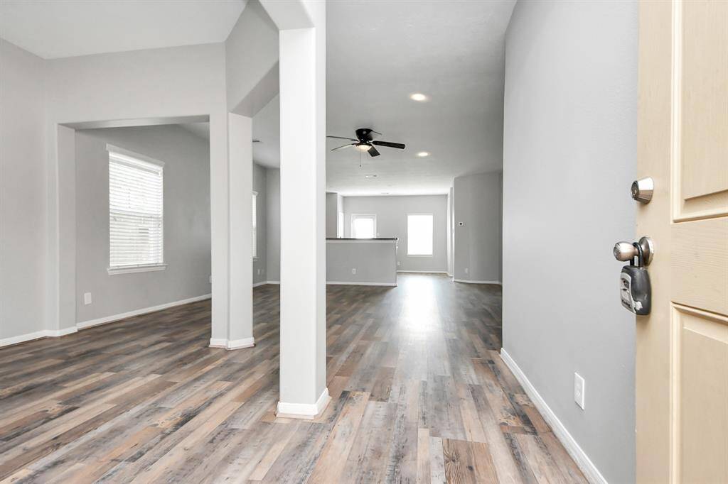 This photo showcases a bright, open living space with modern wood flooring, neutral gray walls, and ample natural light from multiple windows. The area features a ceiling fan and leads into a kitchen with a partial view.