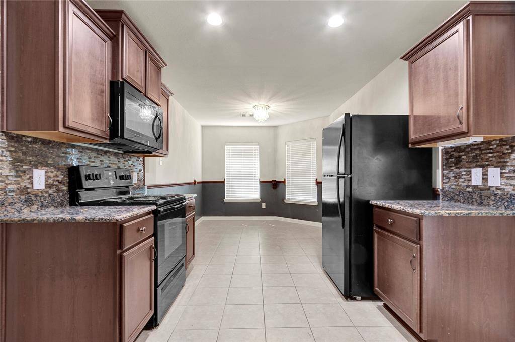 This kitchen features sleek dark appliances, granite countertops, and warm wood cabinetry. The tile flooring complements the mosaic backsplash, and the space flows into a bright dining area with large windows.