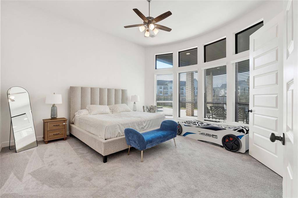 Bright and spacious primary bedroom. The room has a wall of tall windows with blinds, allowing ample natural light, and a ceiling fan for comfort.