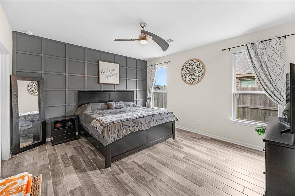 The primary bedroom features a modern aesthetic with a dark custom accent wall and wood-look tile flooring. It includes a ceiling fan, two large windows for natural light, and simple yet stylish decor, creating a cozy and inviting atmosphere.