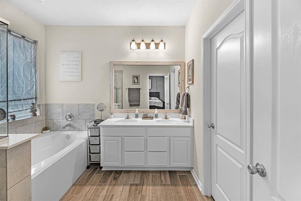 The primary bath features a clean design with a dual-sink vanity, large mirror, and elegant lighting. The space includes a soaking tub with a tiled surround and wood-style tile flooring, offering a clean and inviting atmosphere.