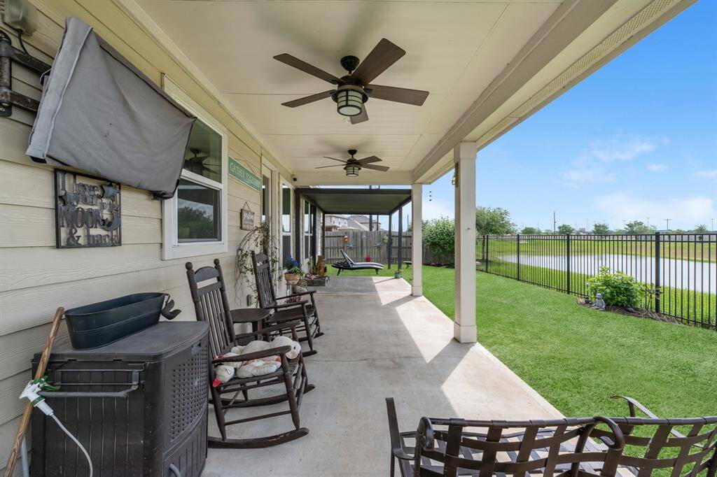 This photo showcases the spacious covered patio with ceiling fans, perfect for relaxation. The patio overlooks a well-maintained lawn and a wrought-iron fenced view of a pond, offering a serene outdoor retreat.  The TV conveys with the sale.