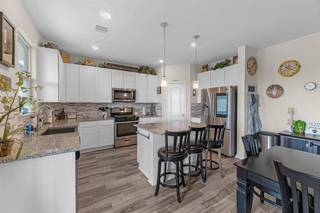 The kitchen features sleek granite countertops, white cabinetry, and stainless-steel appliances. The open layout includes an island with bar seating and elegant pendant lighting. The space connects to a cozy dining area, enhanced by stylish decor and wood-look tile flooring.