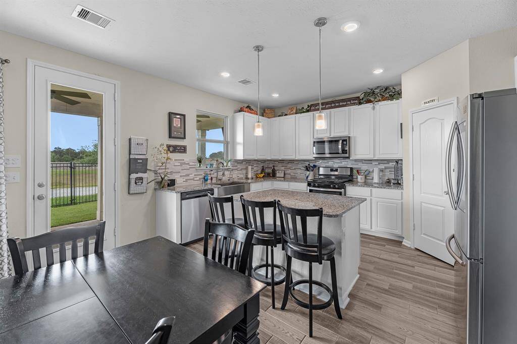 The open layout flows into a dining area with a view of the backyard through a glass door. The space is enhanced by recessed and pendant lighting.