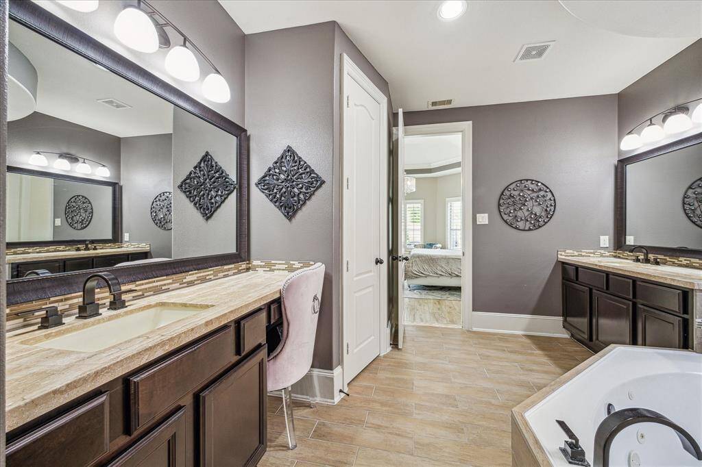 This elegant primary bathroom features separate vanities, granite countertops, framed mirrors, and stylish lighting. The layout, accented by warm wood-look tile flooring, connects seamlessly to the primary bedroom, creating a cohesive and comfortable suite.